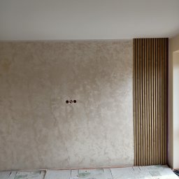 Paweł Stanek - Wykończona ściana z dekoracyjnym tynkiem i drewnianymi lamelami. Widoczne gniazdka elektryczne i zabezpieczona podłoga. Neutralna kolorystyka wnętrza.