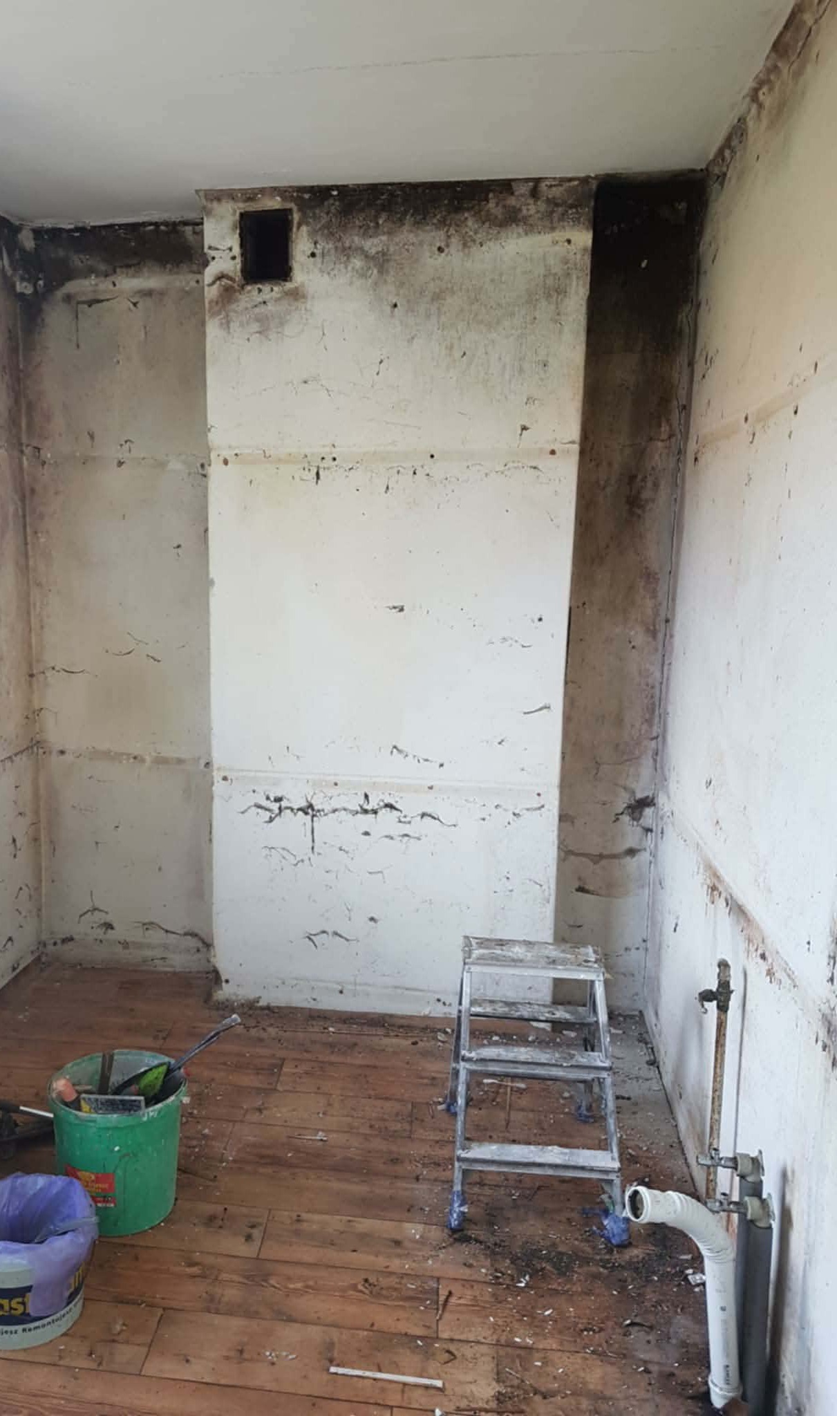 Pokój w trakcie prac przygotowawczych do remontu. Widoczne ślady wilgoci na ścianach, drabina, wiadro z narzędziami i fragment instalacji hydraulicznej.