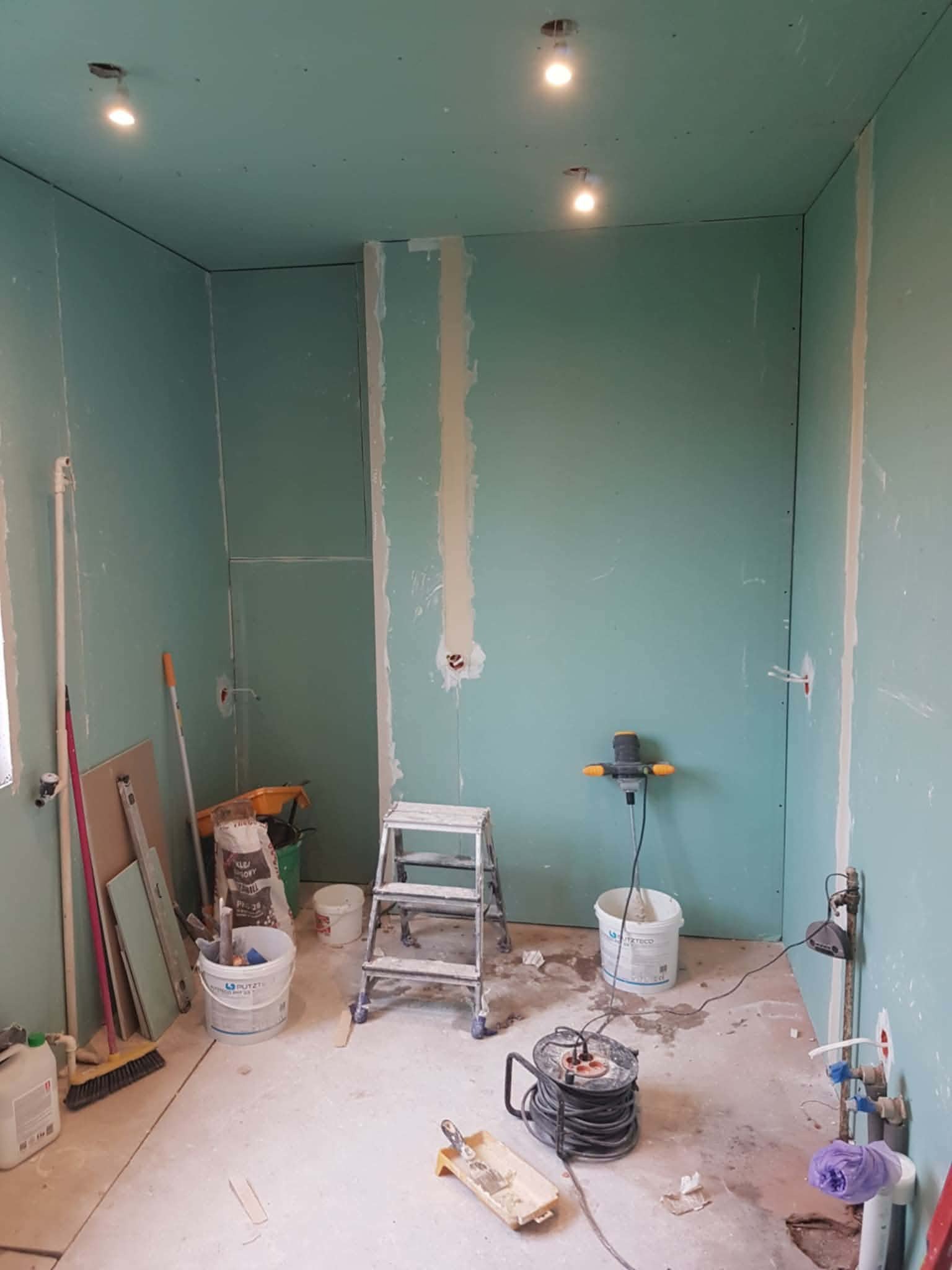 Wnętrze w trakcie montażu płyt kartonowo-gipsowych, widoczne narzędzia, drabina, worki z zaprawą i częściowo zaszpachlowane łączenia płyt. Oświetlenie sufitowe.