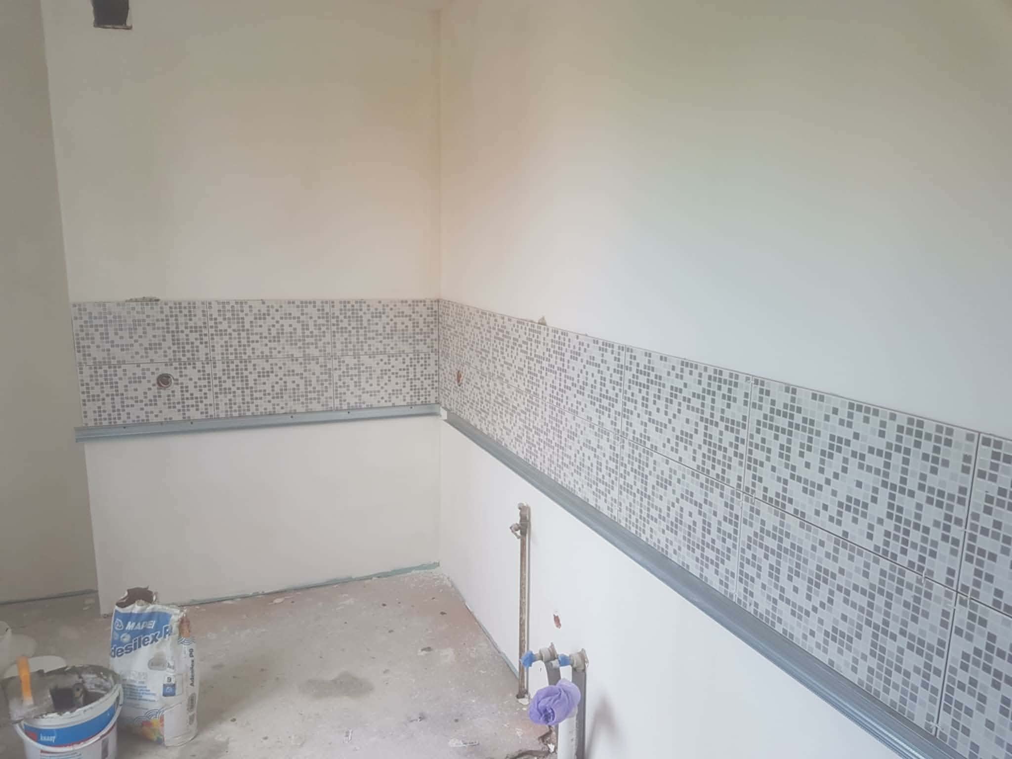 Fragment ściany z mozaiką w trakcie montażu, widoczne profile i materiały budowlane. Prace wykończeniowe w pomieszczeniu z instalacją wodną.