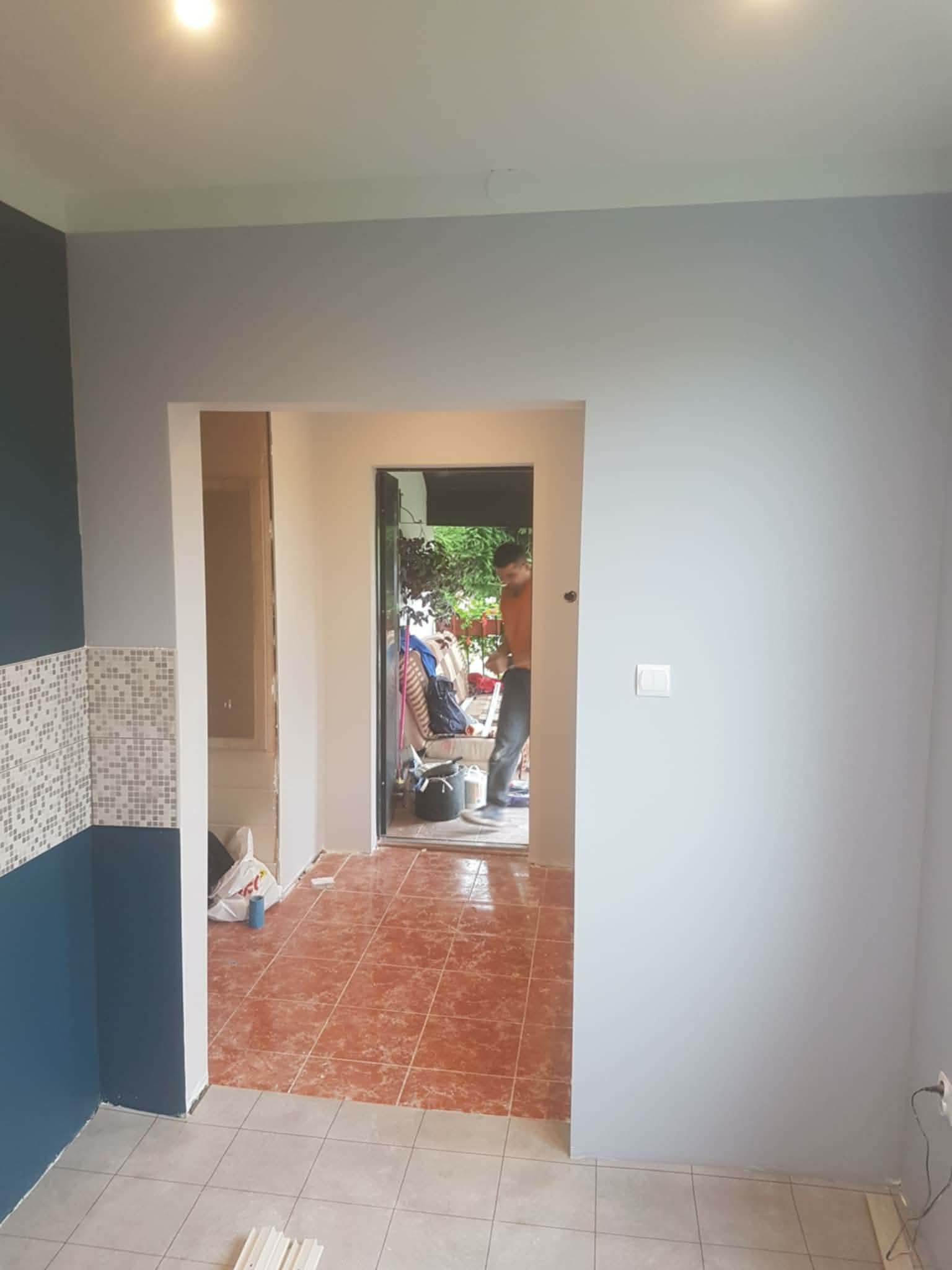 Wnętrze w trakcie odświeżania: pomalowane ściany na szaro i granatowo, fragment mozaiki, widoczny pracownik w przejściu. Podłoga częściowo wyłożona płytkami.