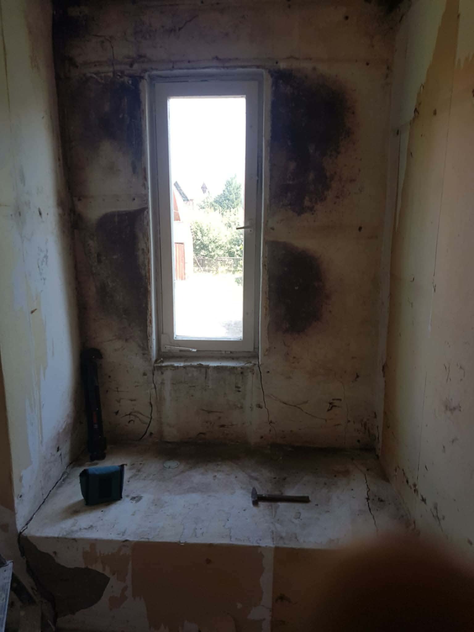 Wnętrze pomieszczenia w trakcie remontu: surowe ściany z widocznymi śladami wilgoci, okno z widokiem na zieleń, narzędzia budowlane na podłodze. Etap przygotowania do prac wykończeniowych.