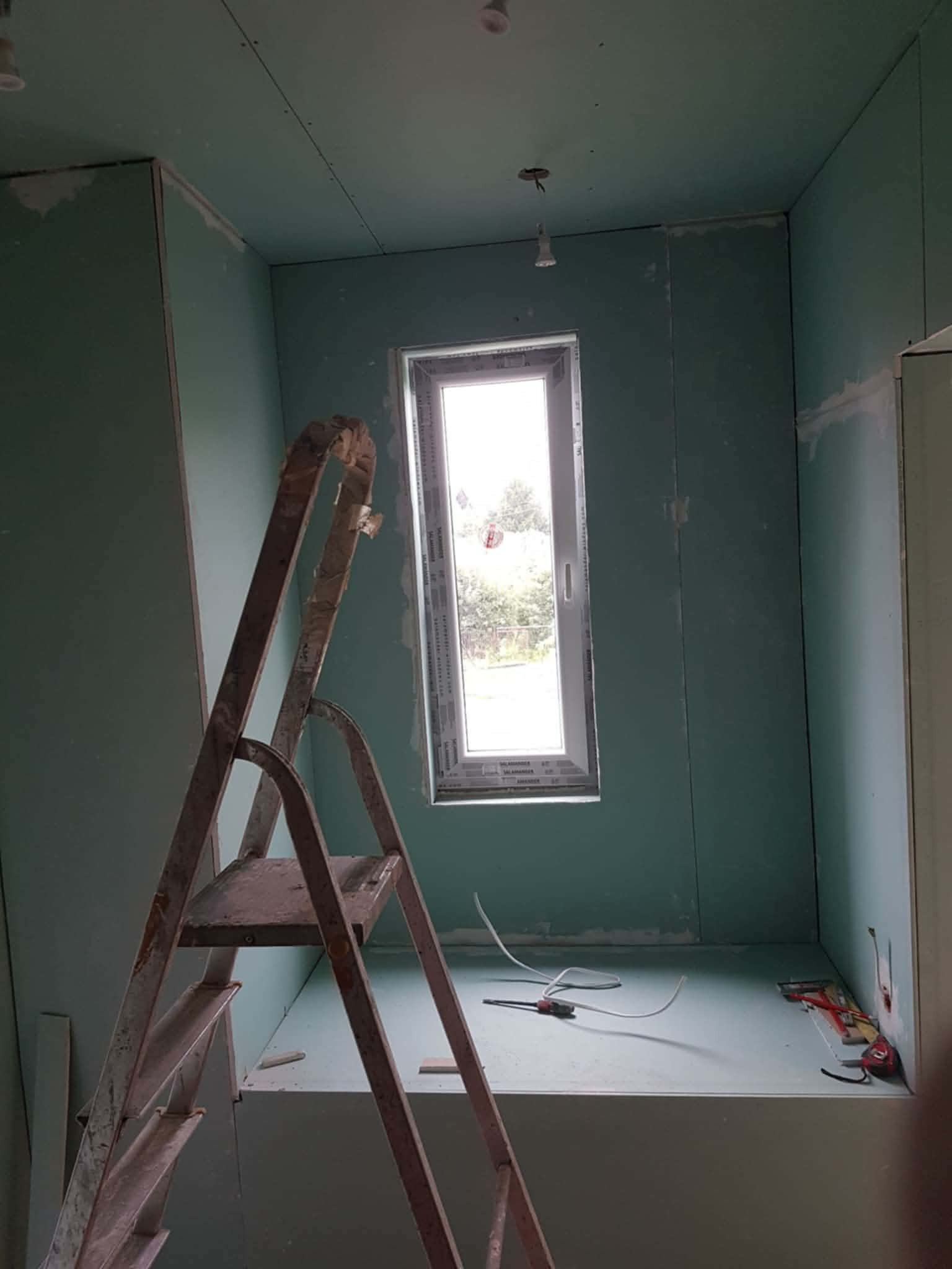 Wnętrze w trakcie montażu płyt gipsowo-kartonowych, widoczna drabina i okno. Ściany przygotowane do dalszych prac wykończeniowych.