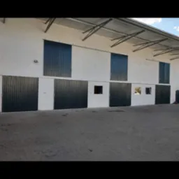 Elewacja budynku z szeregiem ciemnych, przesuwnych bram i okien. Widoczne metalowe elementy konstrukcyjne dachu i betonowa nawierzchnia przed budynkiem. Ujęcie z dołu.