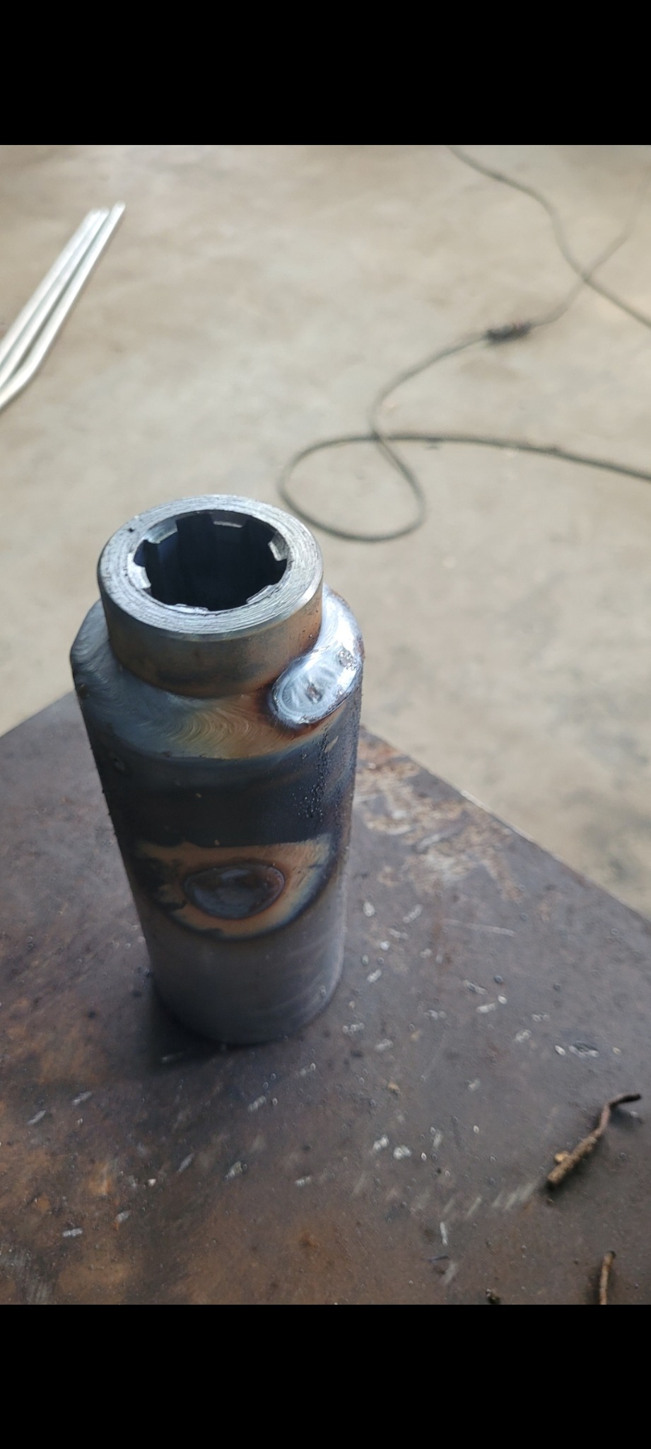 Metalowy cylinder z widocznymi śladami spawania, umieszczony na metalowej powierzchni. Widoczne spawy i obróbka metalu. Detal z bliska.