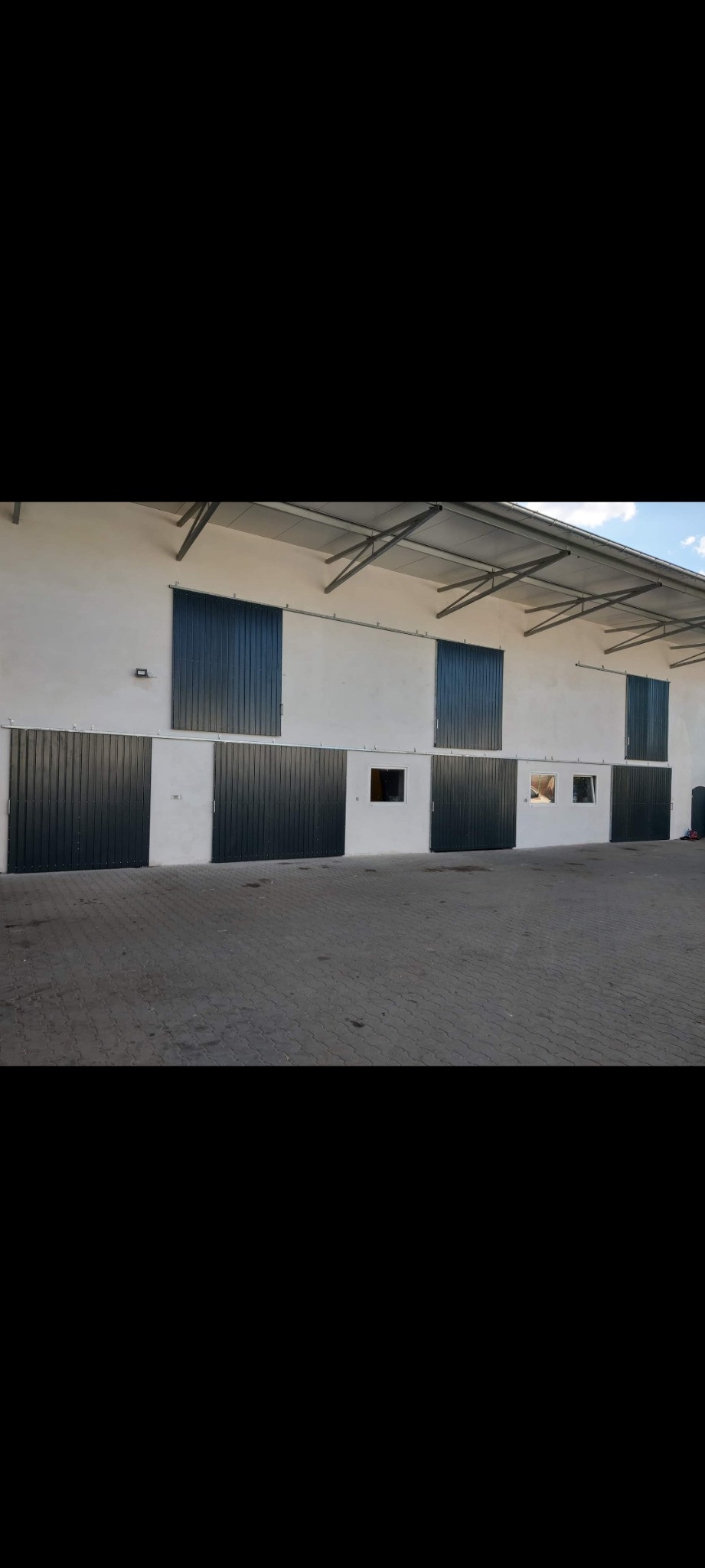 Elewacja budynku z szeregiem ciemnych, przesuwnych bram i okien. Widoczne metalowe elementy konstrukcyjne dachu i betonowa nawierzchnia przed budynkiem. Ujęcie z dołu.