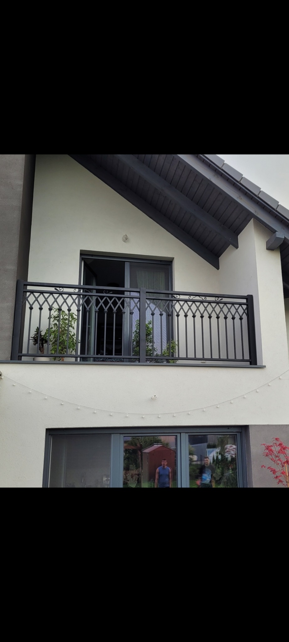 Elegancka, czarna balustrada balkonowa z dekoracyjnym wzorem na tle jasnej elewacji domu. Widoczne rośliny w donicach na balkonie i odbicie osób w oknach.