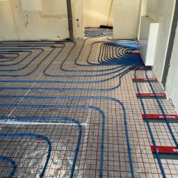 Hydro Instal Tech - Niebieskie rury ogrzewania podłogowego ułożone na folii z siatką w surowym wnętrzu. Widoczny biały rozdzielacz na ścianie i rura pionowa.