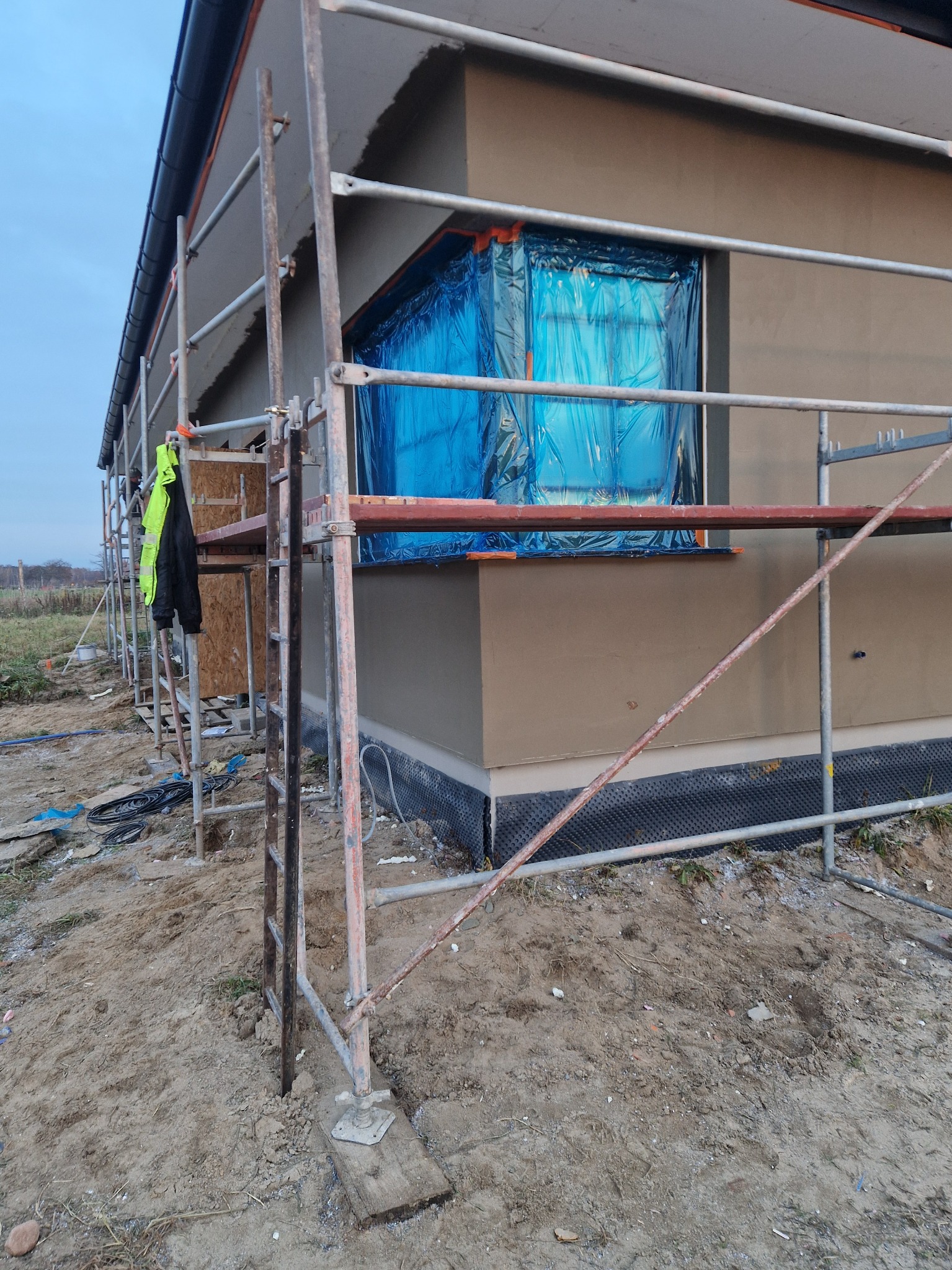 Dom w trakcie tynkowania elewacji, okno zabezpieczone folią, widoczne rusztowanie i teren budowy. Świeży tynk w kolorze beżowym.