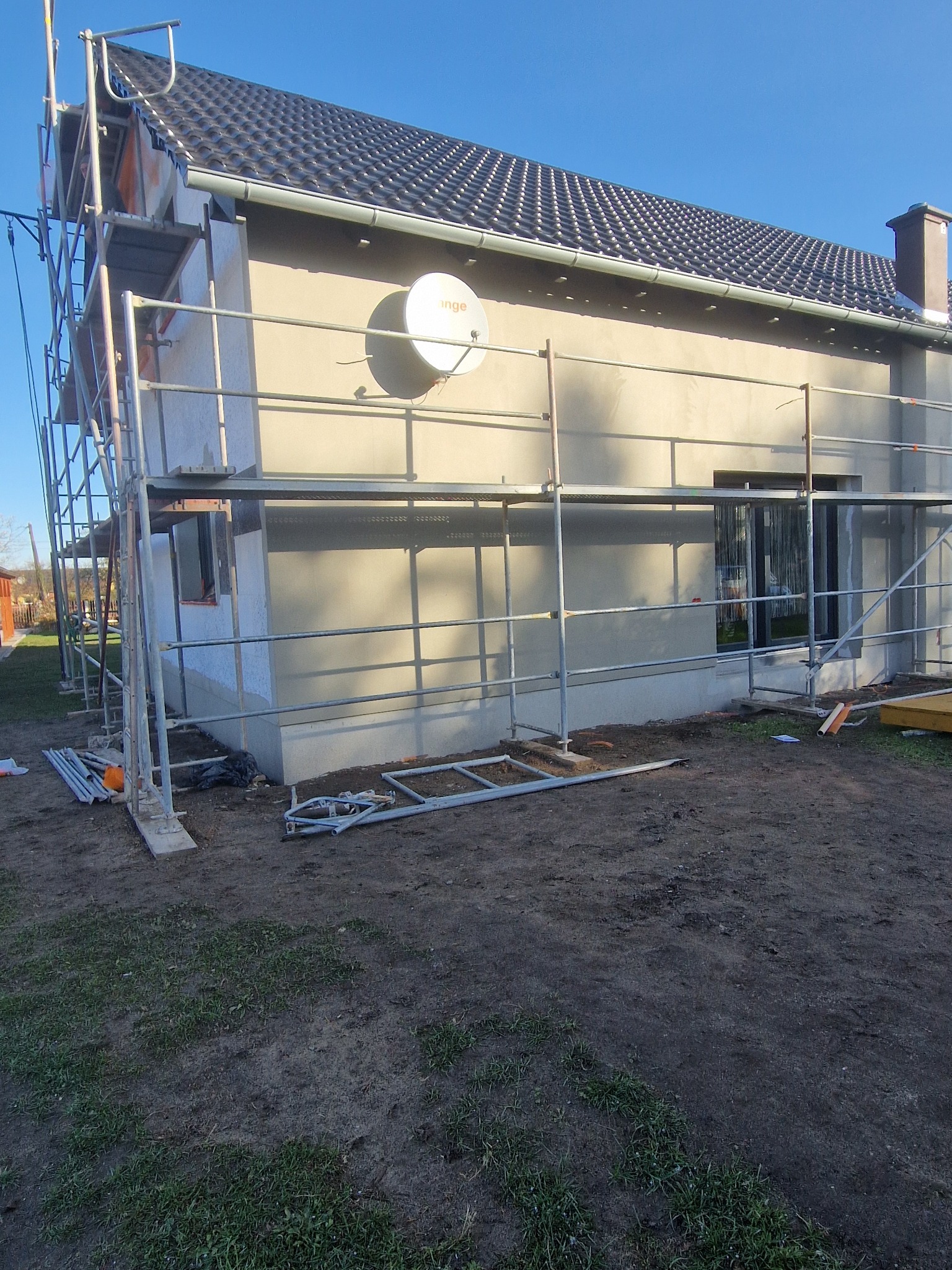 Dom jednorodzinny w trakcie ocieplania elewacji, widoczne rusztowania i częściowo otynkowana ściana. Na ścianie zamontowana antena satelitarna.