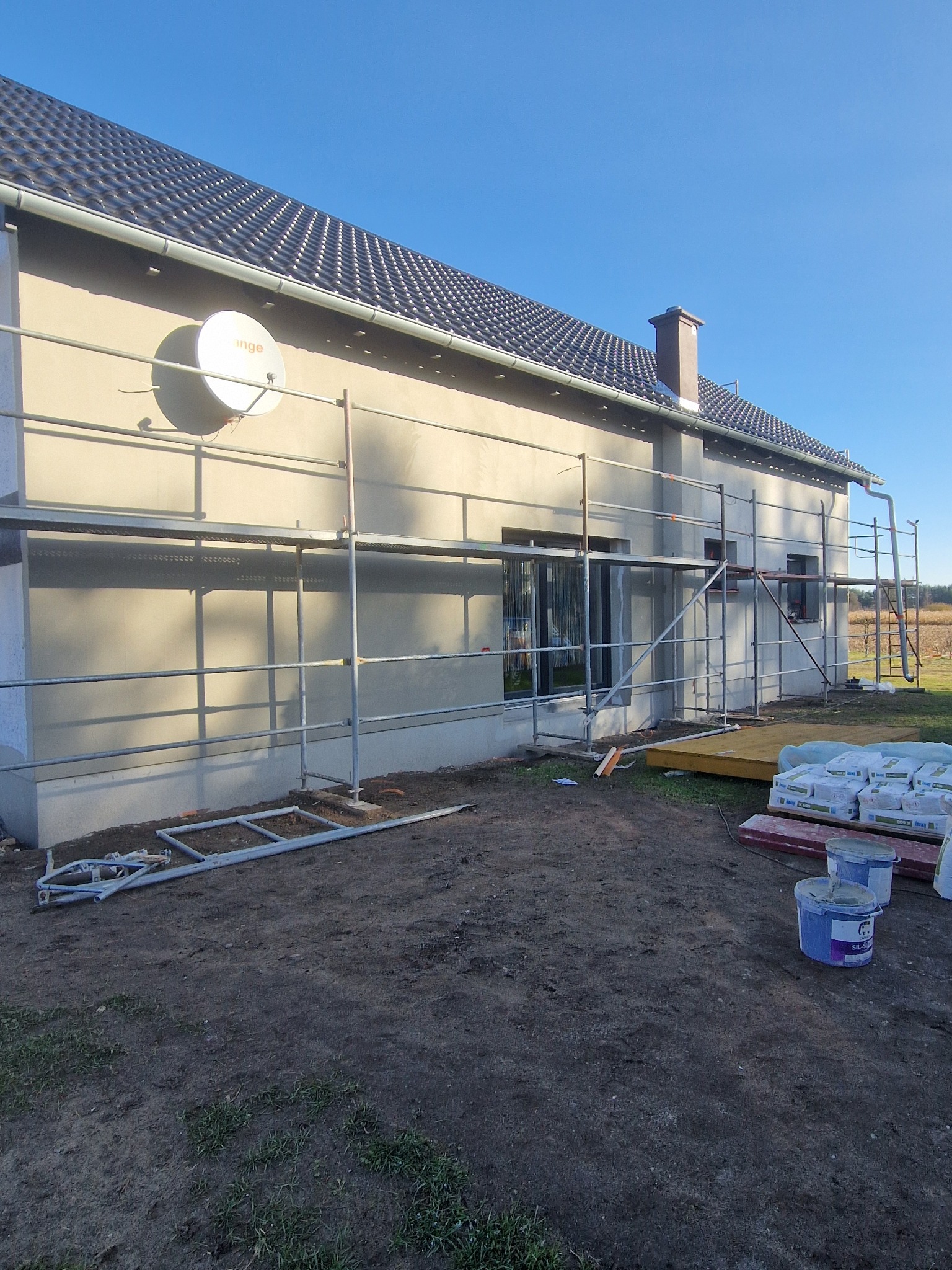 Dom jednorodzinny w trakcie ocieplania, widoczne rusztowania, worki z materiałami budowlanymi i antena satelitarna na elewacji. Słoneczny dzień.