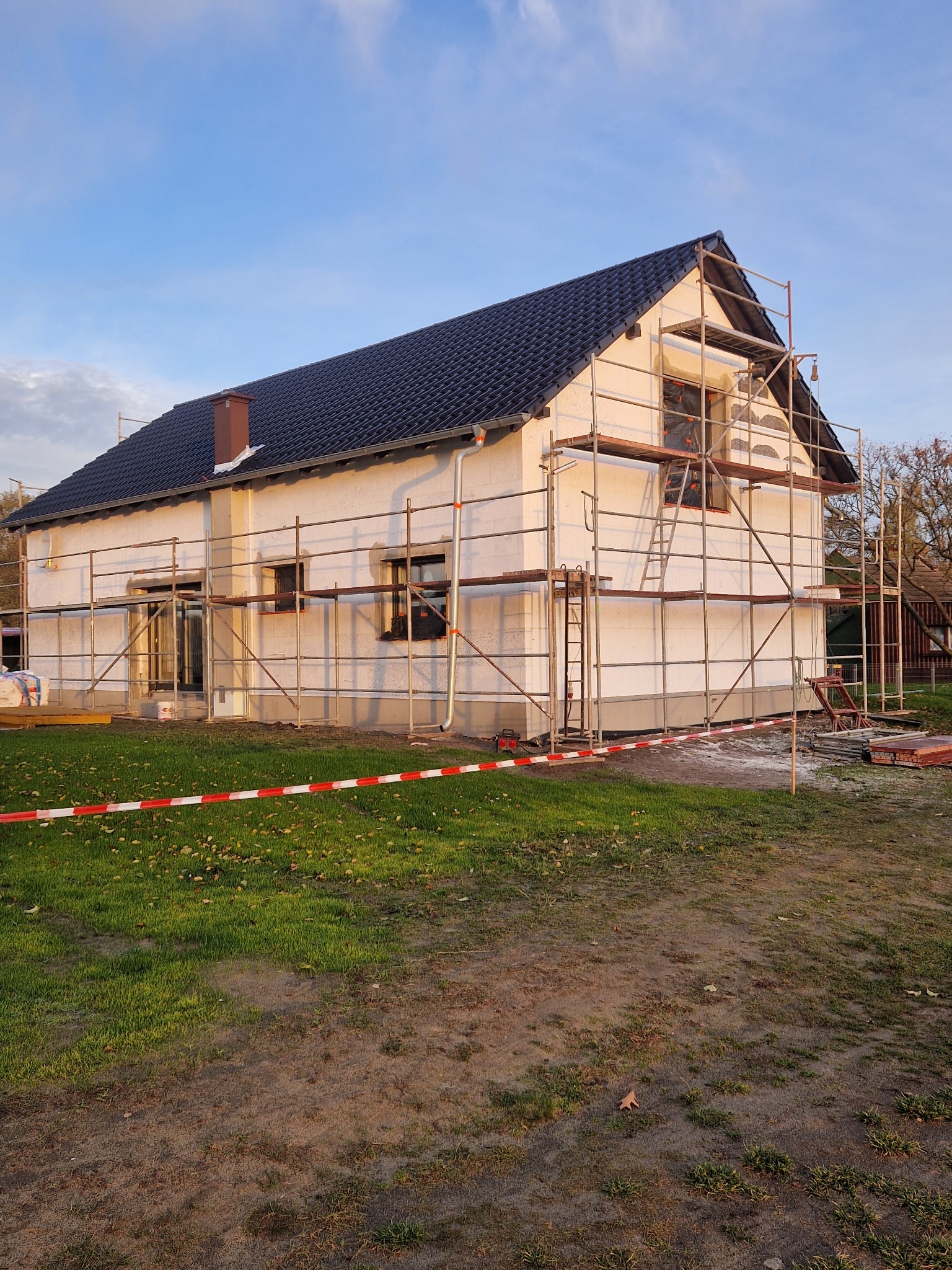 Dom jednorodzinny w trakcie ocieplania, otoczony rusztowaniami i taśmą ostrzegawczą. Widoczne płyty izolacyjne na ścianach i przygotowane materiały budowlane.