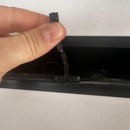 Firma Handlowo-Usługowa Bartosz Lenarcik - Mikro kamera szpiegowska trzymana w palcach, z widocznym obiektywem i taśmą montażową, umieszczona w otworze w ścianie. Instalacja ukrytego monitoringu.