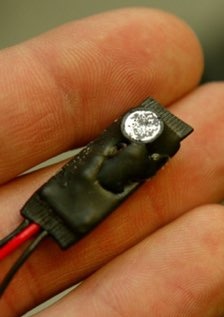 Miniaturowa kamera szpiegowska ukryta w guziku, trzymana w palcach. Widoczne przewody i czarna obudowa. Niewielki rozmiar urządzenia.
