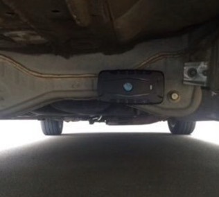 Czarna skrzynka GPS przymocowana pod podwoziem samochodu, widok z dołu, oświetlenie dzienne, koła w tle.