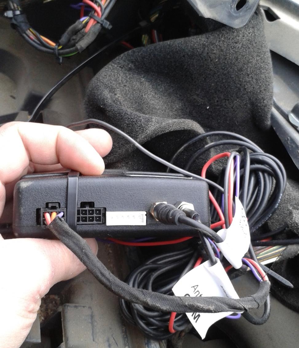 Czarna skrzynka z kablami w dłoni, prawdopodobnie moduł alarmowy lub GPS, z widocznymi złączami i przewodami. Instalacja w pojeździe, wnętrze samochodu.