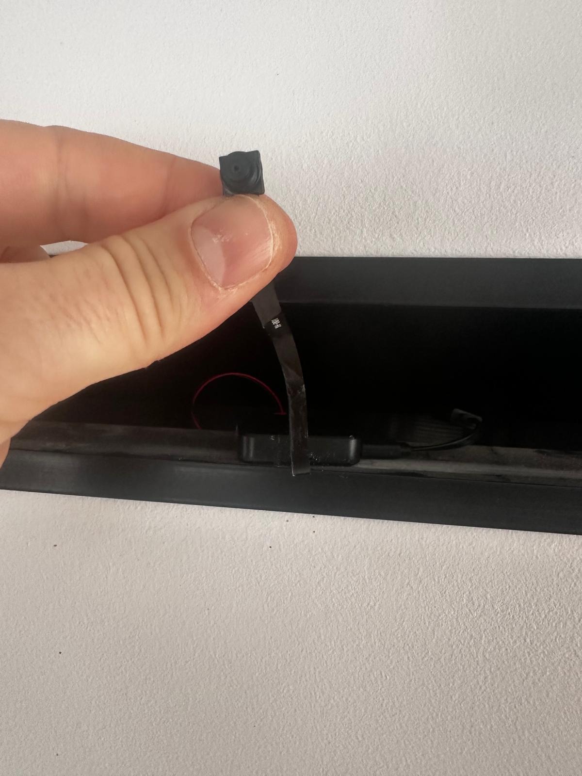Mikro kamera szpiegowska trzymana w palcach, z widocznym obiektywem i taśmą montażową, umieszczona w otworze w ścianie. Instalacja ukrytego monitoringu.