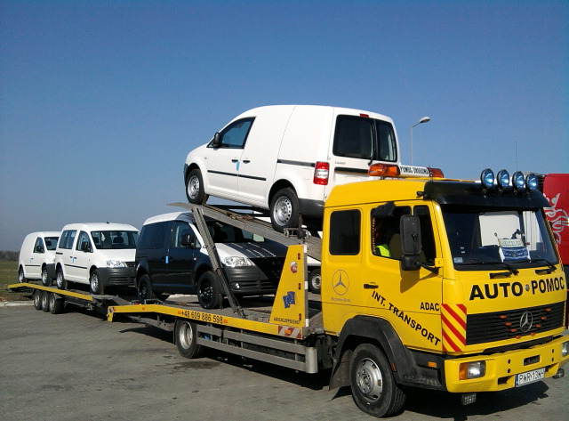 Żółta laweta Mercedes-Benz z trzema samochodami typu van na platformie, w tym jeden biały na górze. Widoczne logo 'Auto Pomoc' i oznaczenia 'Int. Transport'.