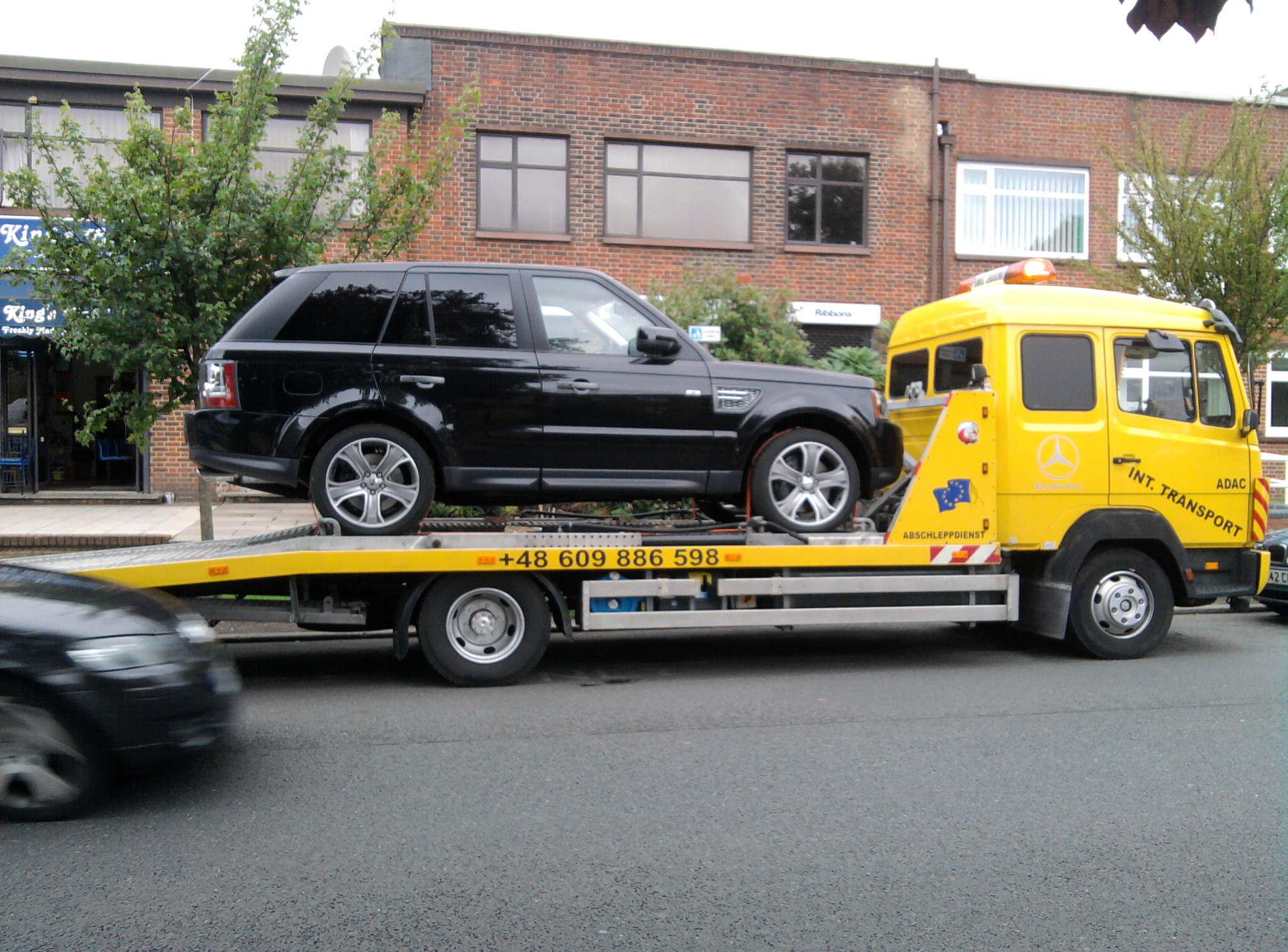 Żółta laweta z czarnym SUV-em na platformie, widoczny numer telefonu. W tle budynek z cegły i zieleń. Usługa transportu pojazdów.