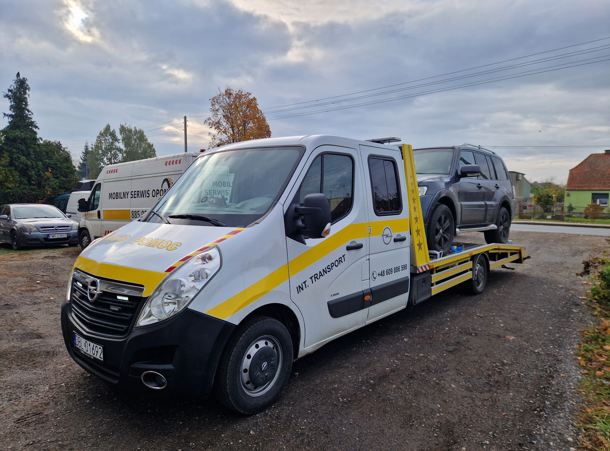 Laweta Opel z szarym SUV-em na platformie, zaparkowana na żwirowym placu. Widoczny numer telefonu i napis 'INT. TRANSPORT'. W tle inne pojazdy i budynki.