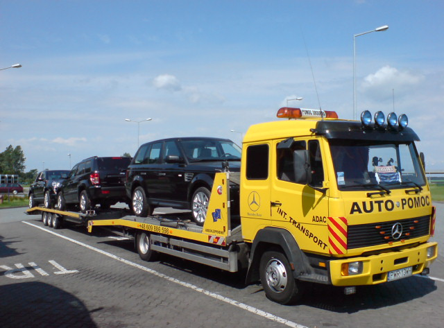Żółta laweta Mercedes-Benz z napisem 'AUTO-POMOC' przewozi trzy czarne samochody Range Rover na platformie. Widoczne numery telefonu i logo ADAC.