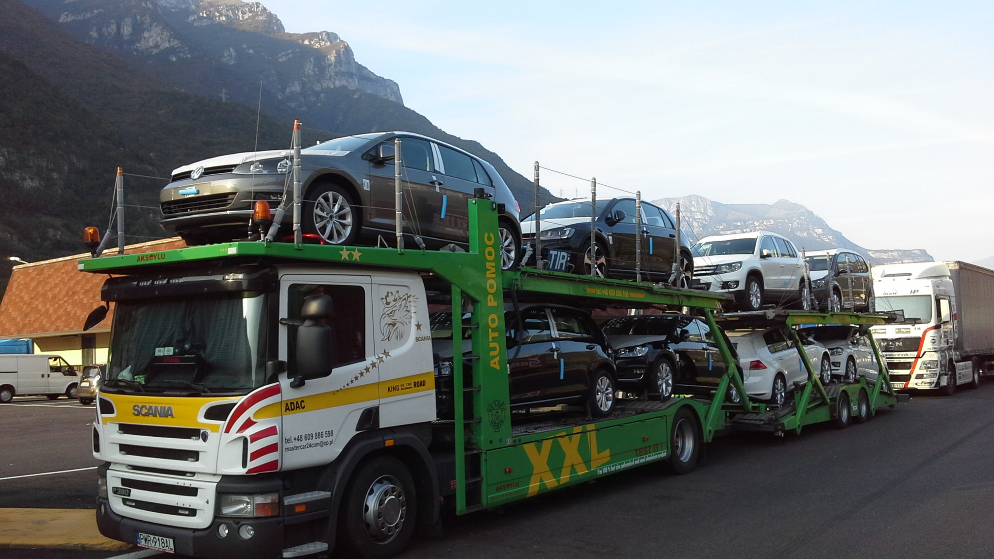 Laweta Scania z załadowanymi samochodami VW na tle gór. Widoczny numer telefonu i logo ADAC. Transport aut z zagranicy.