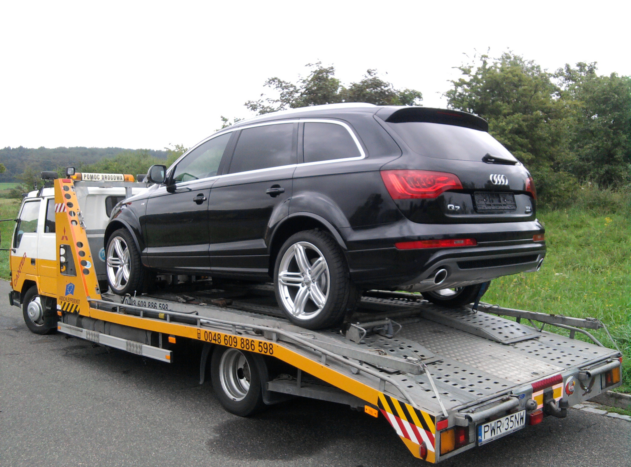 Czarny SUV Audi Q7 na żółto-białej lawecie pomocy drogowej z numerem telefonu. Widok z tyłu, tablica rejestracyjna widoczna. Trawa i drzewa w tle.