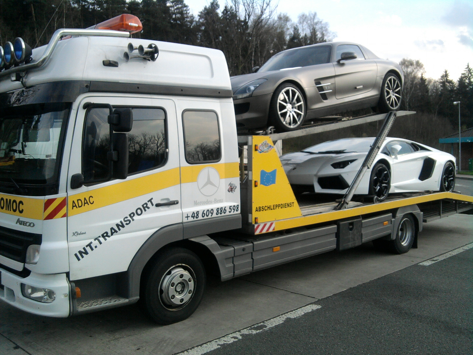 Laweta z dwoma luksusowymi autami: srebrnym Mercedesem SLS AMG na górze i białym Lamborghini Aventador na dole. Żółto-biała laweta z napisem 'INT.TRANSPORT'.