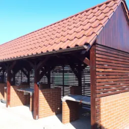 Zadaszona, drewniana konstrukcja z ceglanymi elementami i brązową dachówką ceramiczną w słoneczny dzień. Widoczne rynny i elementy wykończeniowe.