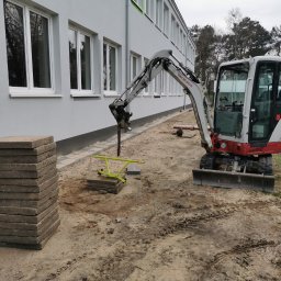 VOLT DAMIAN POLAŃSKI - Mini koparka Takeuchi przygotowuje teren pod ułożenie kostki brukowej. Stos kostki obok wykopu. Budynek w tle. Widoczne ślady opon na piasku.