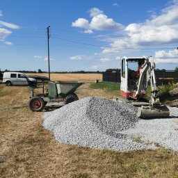 VOLT DAMIAN POLAŃSKI - Mini koparka i wywrotka obok pryzmy żwiru na placu budowy. W tle budynek, samochody i pole pod błękitnym niebem z chmurami.