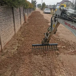 Mini koparka Takeuchi z grabiami wyrównuje teren wzdłuż ogrodzenia z betonowych płyt. W tle droga, samochód i zabudowania. Pochmurne niebo.
