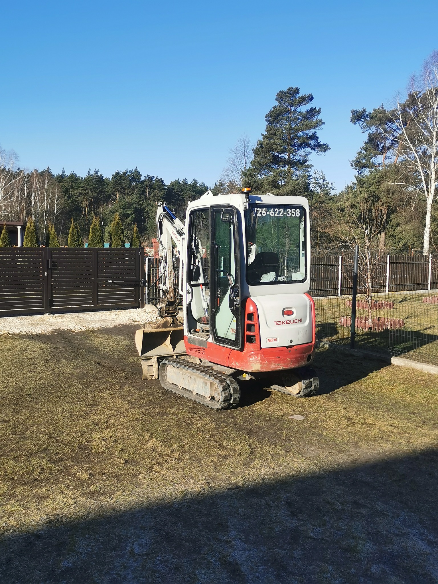 Kompaktowa minikoparka Takeuchi TB216 na trawiastym terenie, z widocznym numerem telefonu na kabinie, sugerująca usługi wykopów lub niwelacji terenu.
