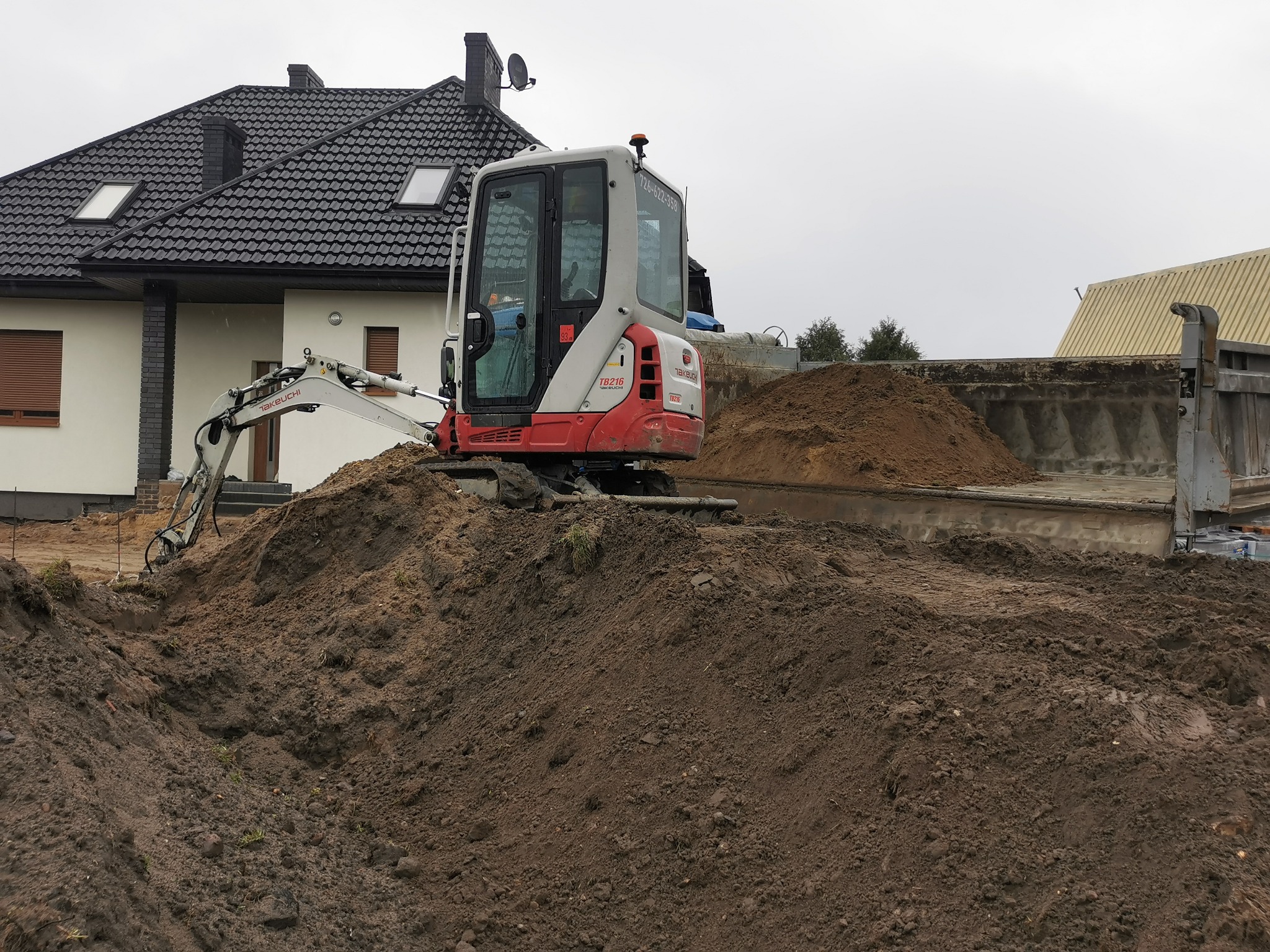 Mini koparka Takeuchi na hałdzie ziemi, obok wywrotka. Prace ziemne w Krzyżanowie. Widoczny dom z ciemną dachówką i fragment budynku gospodarczego.