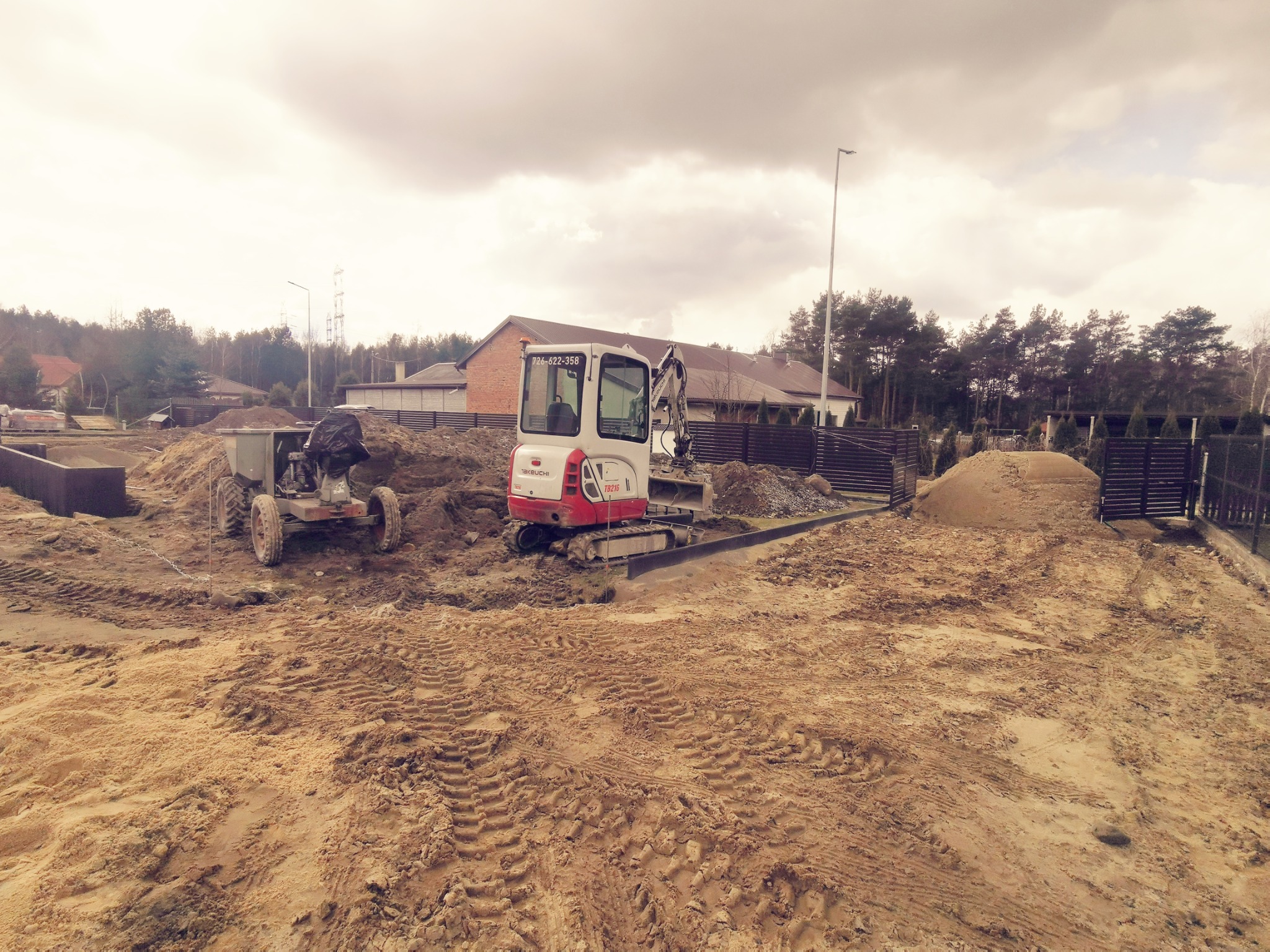 Mini koparka Takeuchi TB216 na placu budowy, obok przyczepa z ziemią. Widoczne ślady opon na piasku i elementy ogrodzenia w tle. Pochmurne niebo.