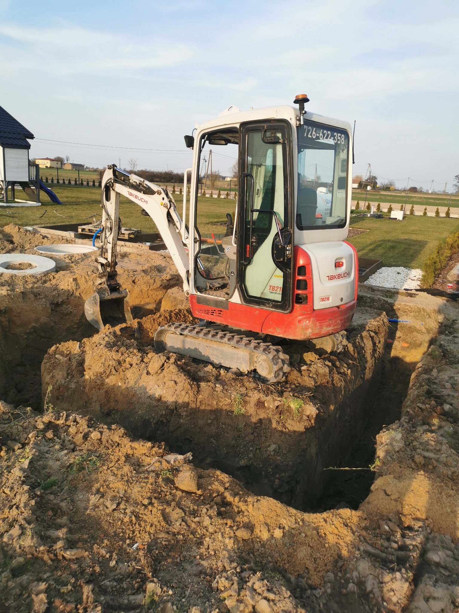 Mini koparka Takeuchi TB216 w trakcie wykopu rowu na działce z elementami szamba. Widoczne betonowe kręgi i świeżo wykopana ziemia. Słoneczny dzień.