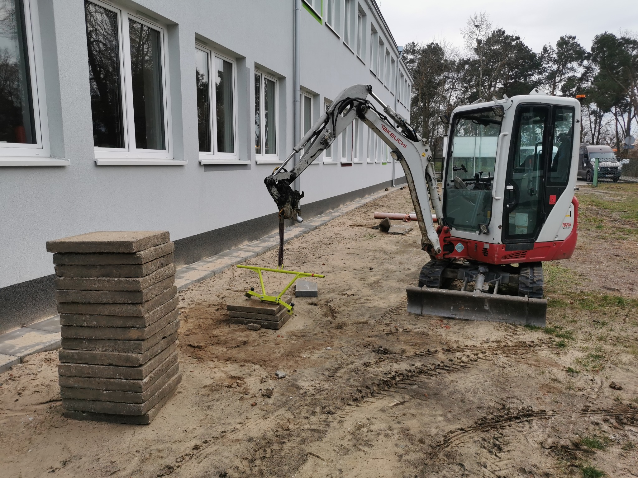 Mini koparka Takeuchi przygotowuje teren pod ułożenie kostki brukowej. Stos kostki obok wykopu. Budynek w tle. Widoczne ślady opon na piasku.