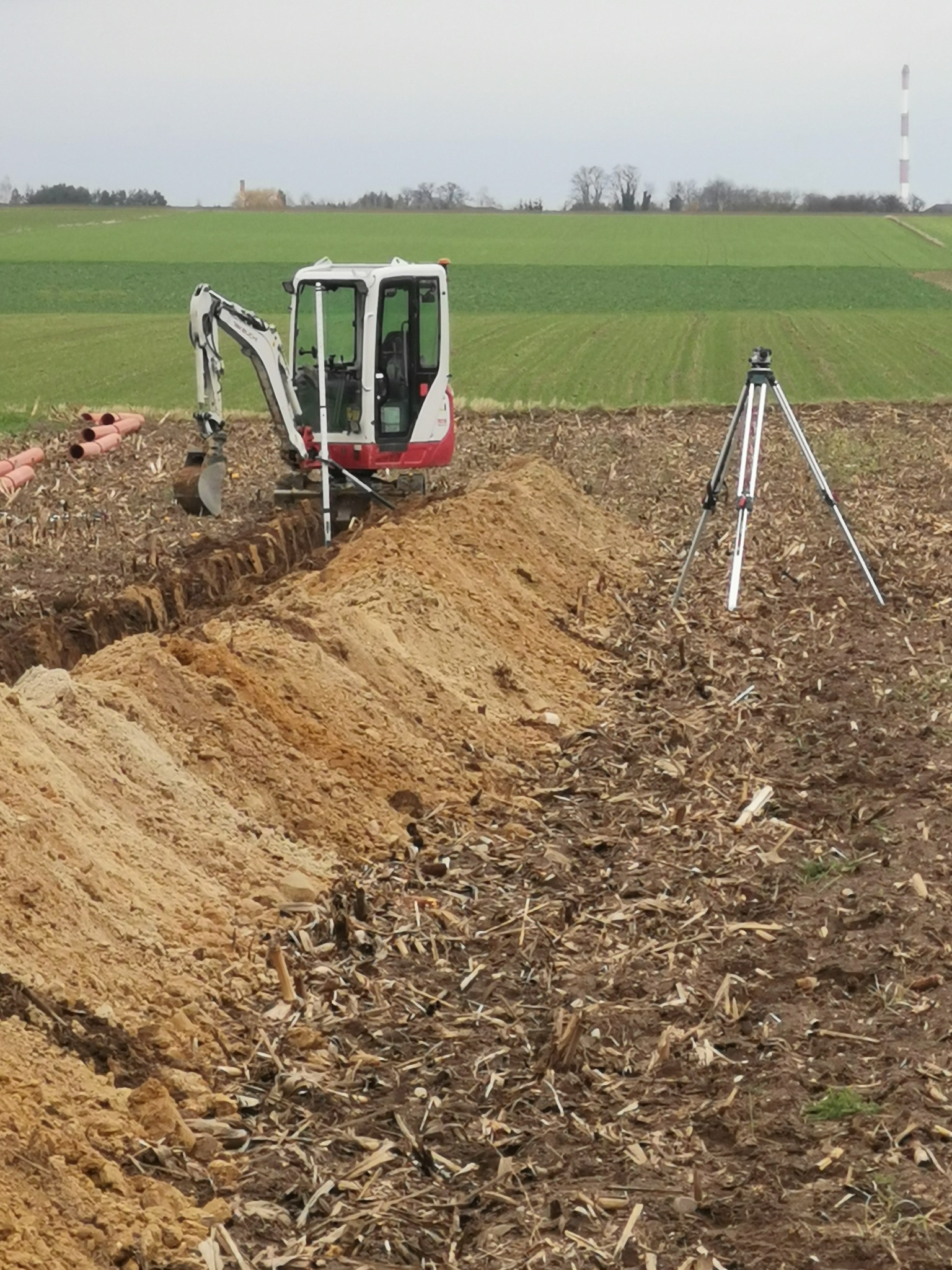 Mini koparka w trakcie wykonywania wykopu pod rury na polu. W tle widoczne zielone pola i komin. Na pierwszym planie statyw geodezyjny.
