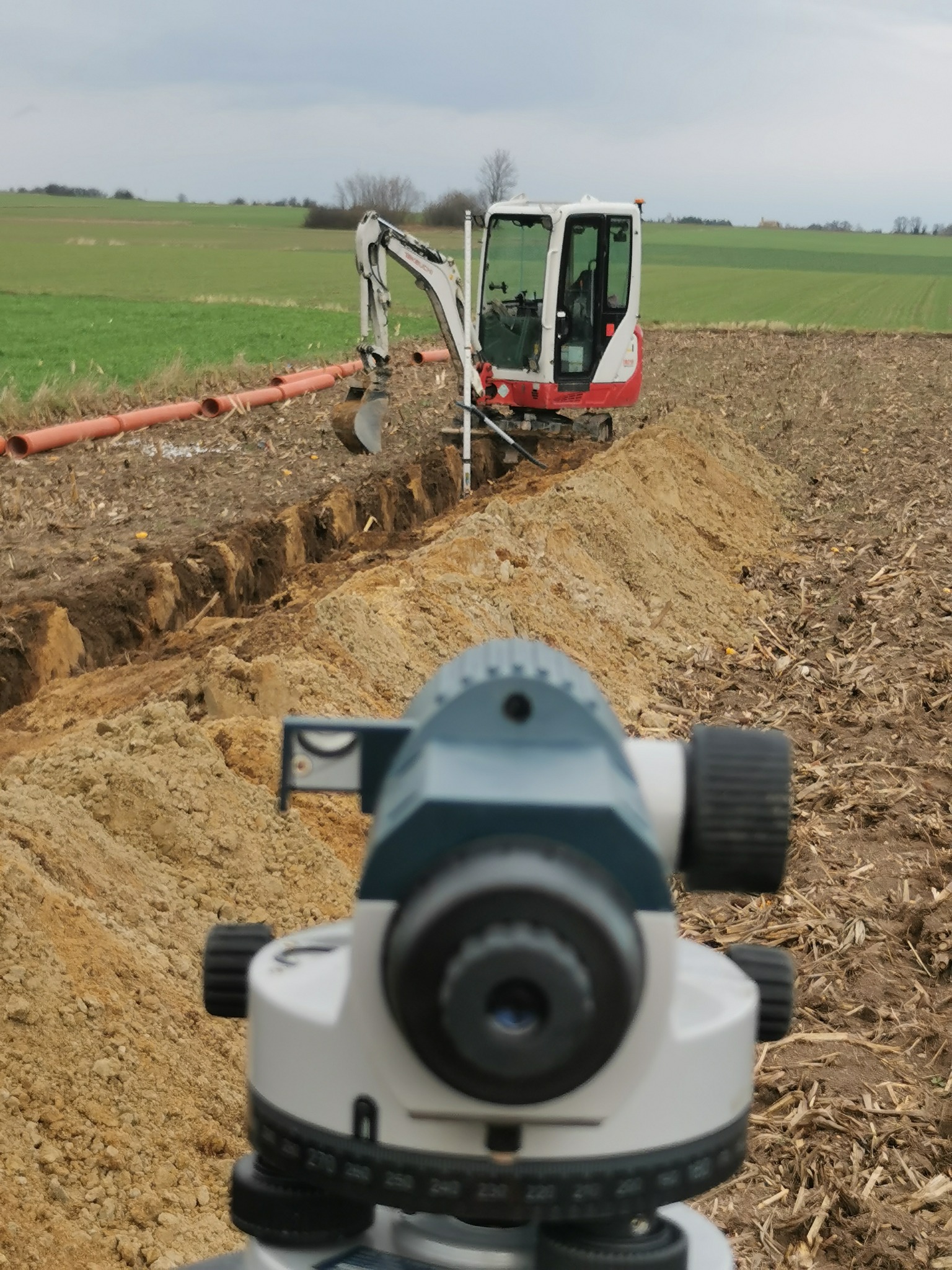 Niwelator na pierwszym planie, w tle minikoparka przy wykopie pod rury na polu. Prace ziemne w toku, pochmurne niebo.