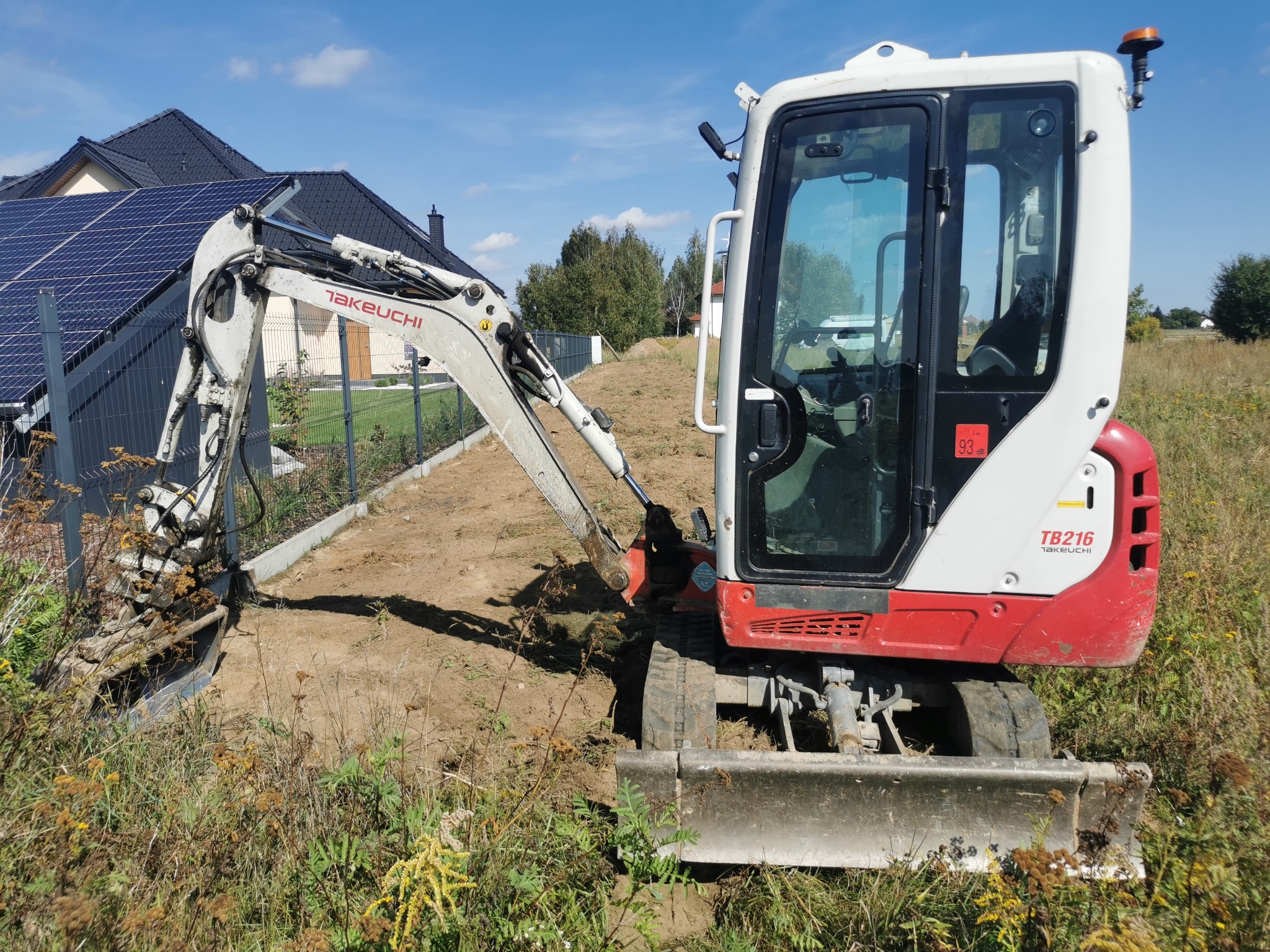 Mini koparka Takeuchi TB216 na działce z domem i panelami słonecznymi w tle. Wykop pod ogrodzeniem, widoczne ślady pracy. Słoneczny dzień.