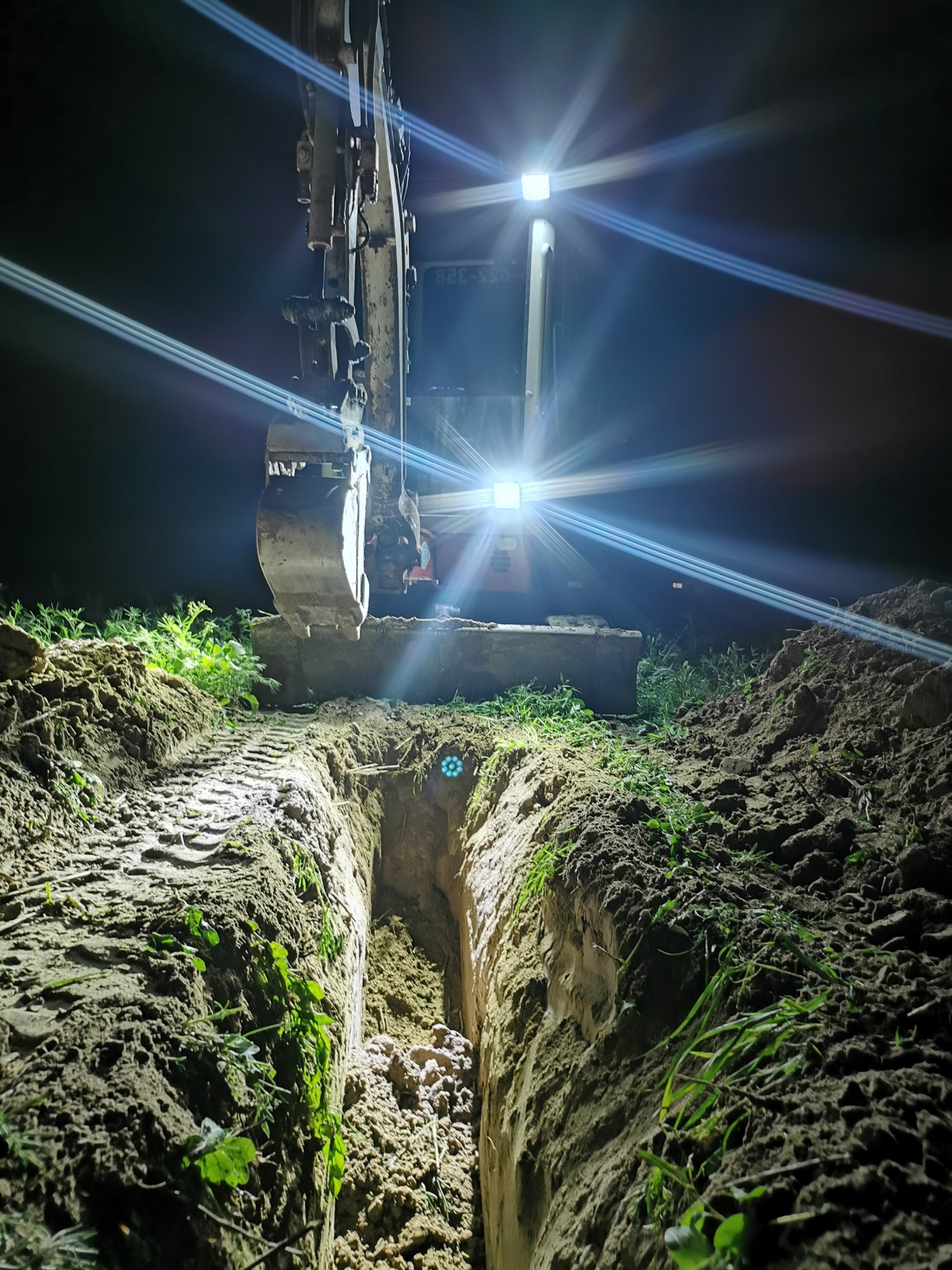 Wykop wąskoprzestrzenny oświetlony reflektorami koparki w nocy. Ślady opon na ziemi, świeża zieleń. Ujęcie z dołu, perspektywa zagłębiająca się w wykop.
