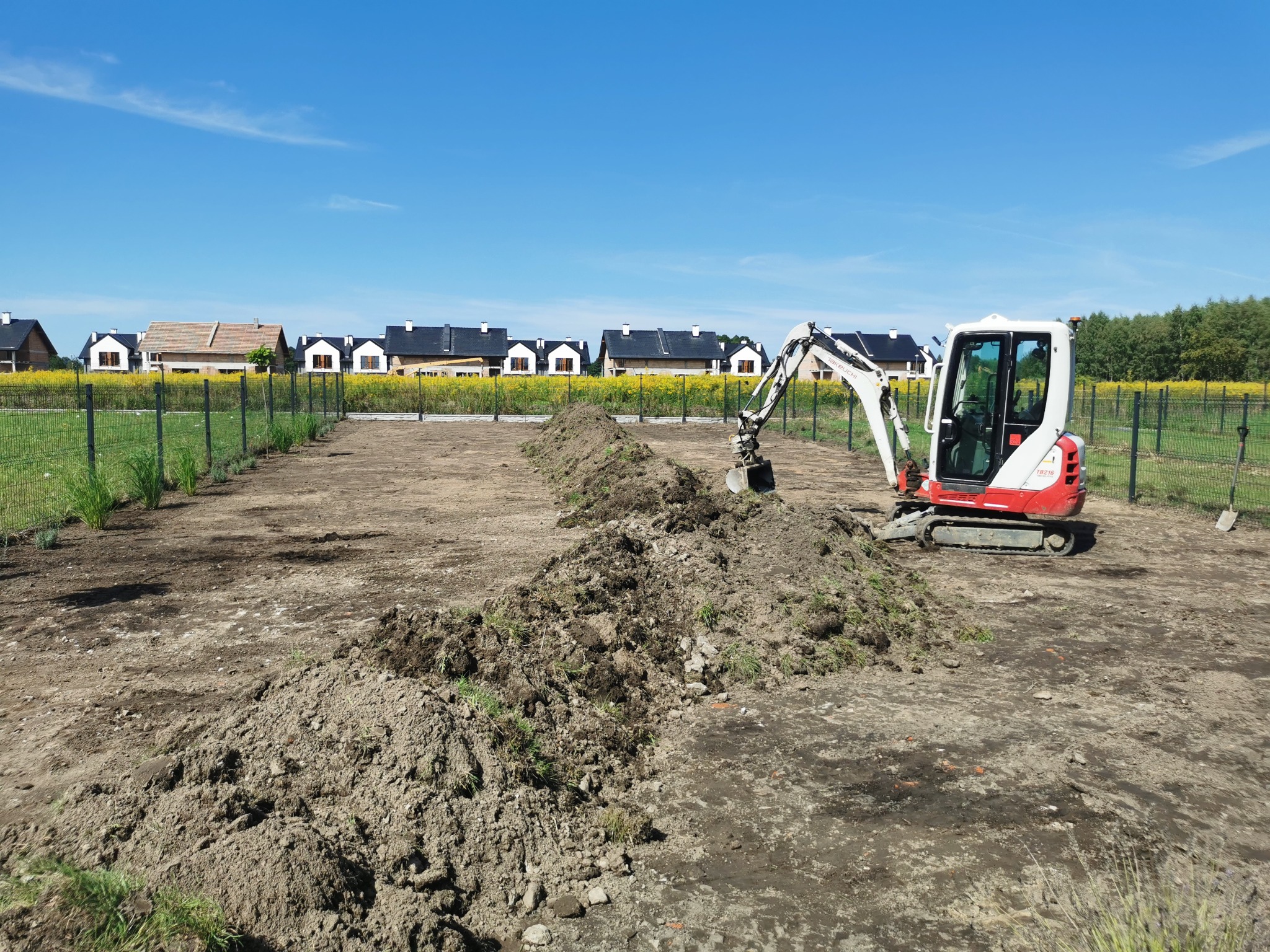 Czerwono-biała mini koparka Takeuchi na działce z pryzmą ziemi, w tle ogrodzenie i rząd domów jednorodzinnych pod błękitnym niebem.