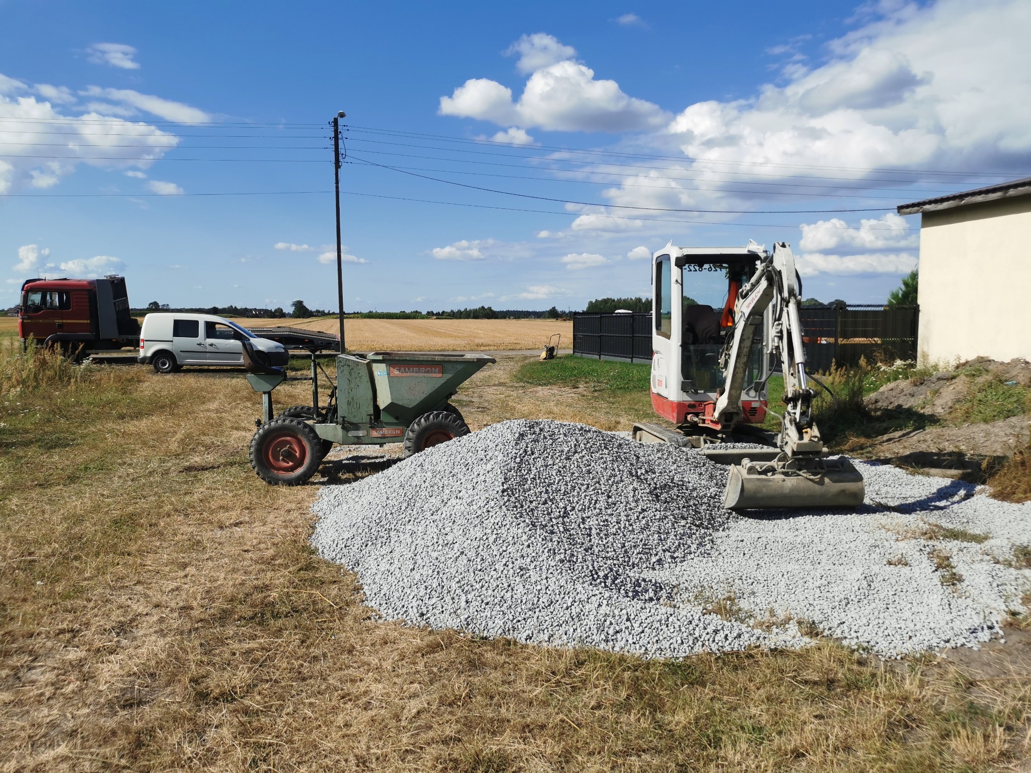 Mini koparka i wywrotka obok pryzmy żwiru na placu budowy. W tle budynek, samochody i pole pod błękitnym niebem z chmurami.