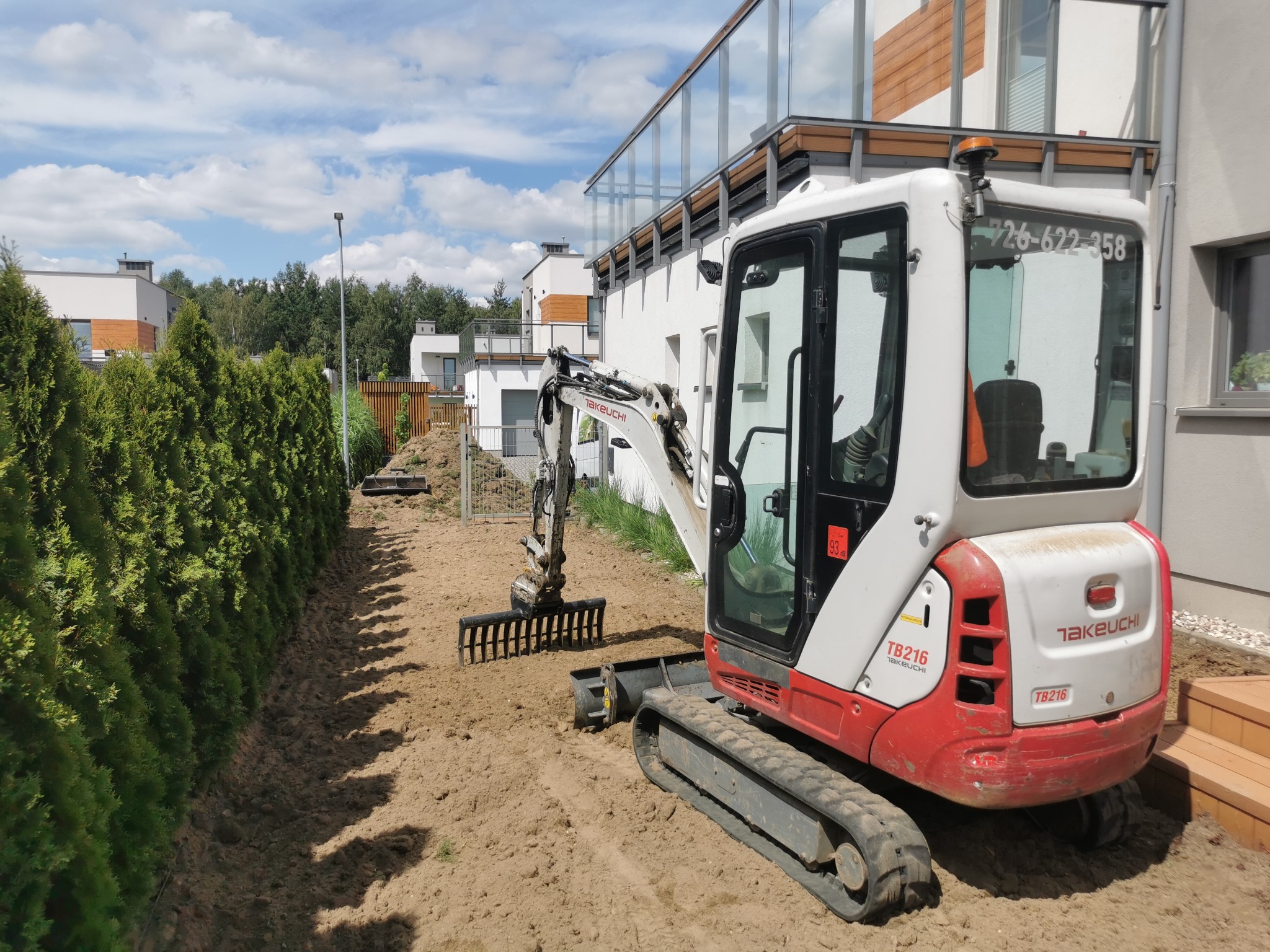 Mini koparka Takeuchi TB216 na działce, przygotowuje teren pod nasadzenia. Widoczne świeżo wyrównane podłoże i rząd tuj. Nowoczesne budynki w tle.