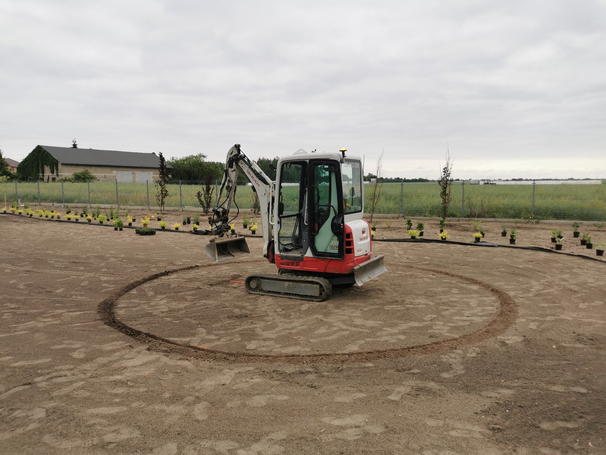 Mini koparka Takeuchi TB216 w trakcie prac ziemnych, wykop w kształcie okręgu, przygotowanie terenu pod nasadzenia. Widok na budynek w tle.