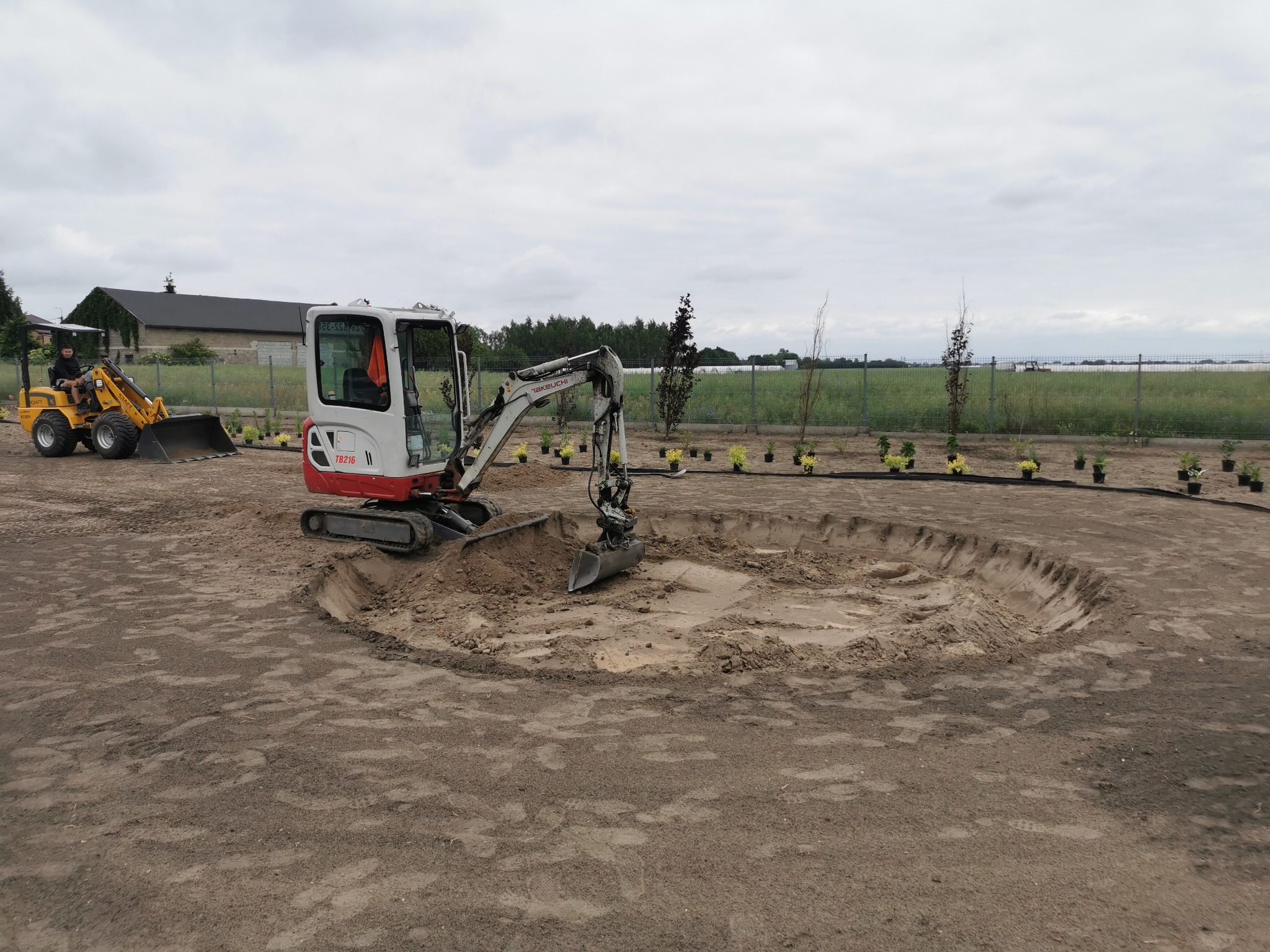 Mini koparka Takeuchi TB216 w trakcie wykopu okrągłego stawu na działce. W tle widoczny operator na małej ładowarce i przygotowane sadzonki drzew.