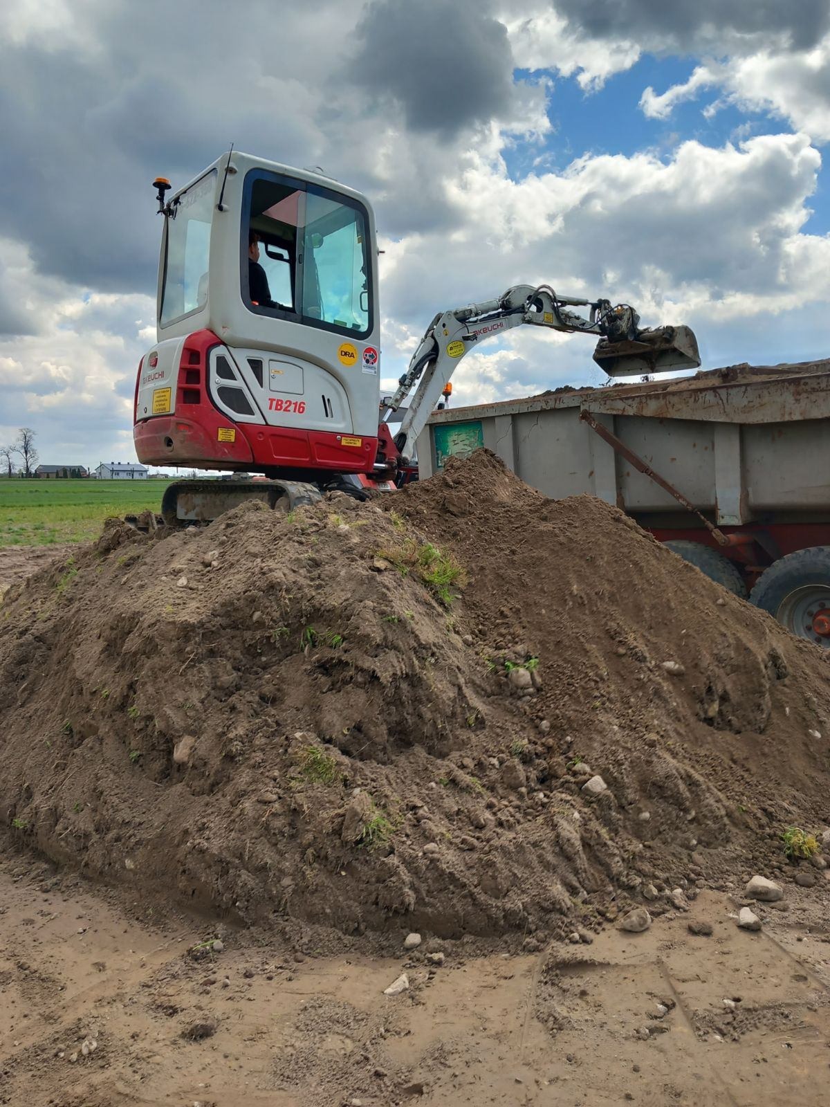 Mini koparka Takeuchi TB216 nabiera ziemię z hałdy i wrzuca do przyczepy. Widoczne pole w tle i pochmurne niebo. Praca w toku, maszyna w akcji.