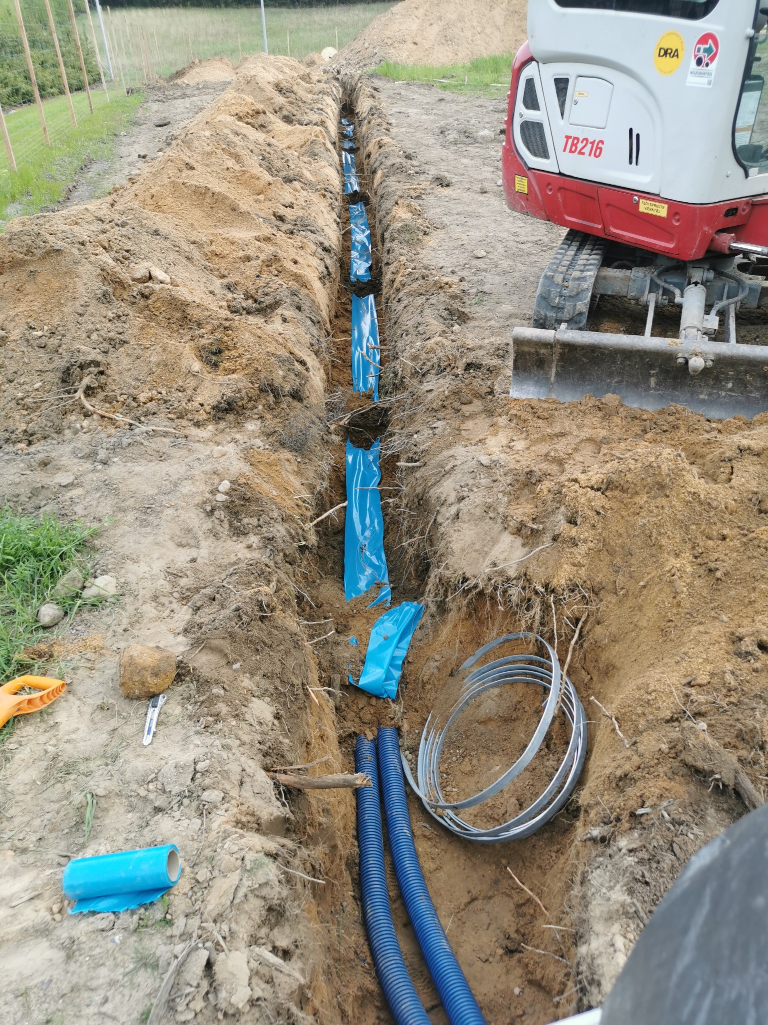 Wykop z niebieskimi rurami i folią ochronną, widoczny fragment koparki. Prace ziemne w toku, przygotowanie pod instalacje. Zabezpieczenie rur przed uszkodzeniem.