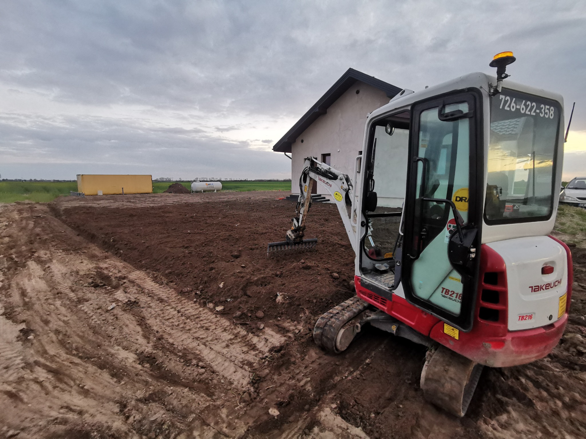 Mini koparka Takeuchi TB216 na placu budowy, przygotowuje teren pod budowę domu. Widoczny wykopany rów i budynek w tle. Numer kontaktowy na kabinie.
