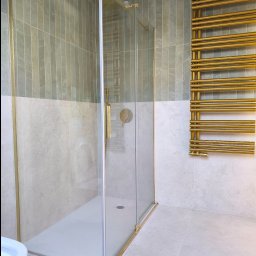 Dawid Radzewicz - Nowoczesna kabina prysznicowa z złotymi detalami, grzejnik drabinkowy w kolorze złota, płytki w odcieniach szarości. Minimalistyczny design łazienki.